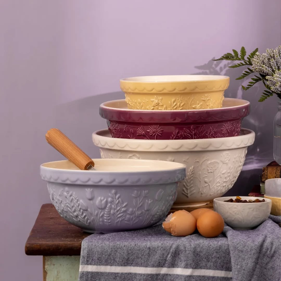 Stack of Mason Cash embossed ceramic mixing bowls in lavender, cream, berry, and yellow, styled with eggs and a wooden pastry brush on a rustic wooden table
