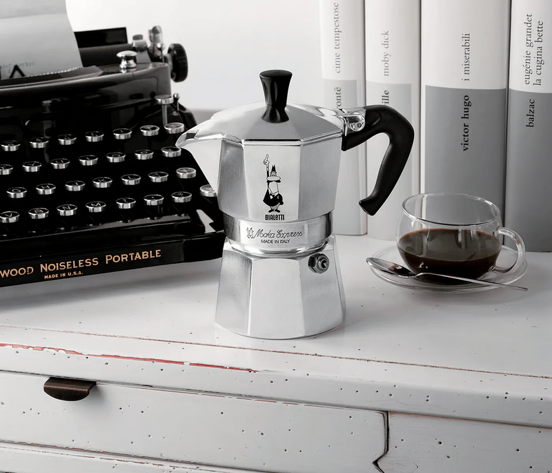 Bialetti Moka Express stovetop coffee maker beside a cup of espresso and vintage typewriter