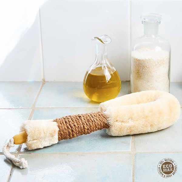 A natural fiber bath scrubber with a beige plush loop handle and a brown rope-wrapped grip, displayed on a light blue tiled surface next to a glass bottle of golden liquid and a clear bottle of white scrub.