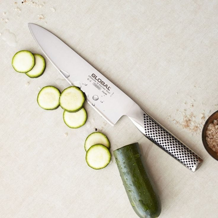 Global stainless steel chef's knife beside freshly sliced courgette rounds and a bowl of sea salt on a stone bench