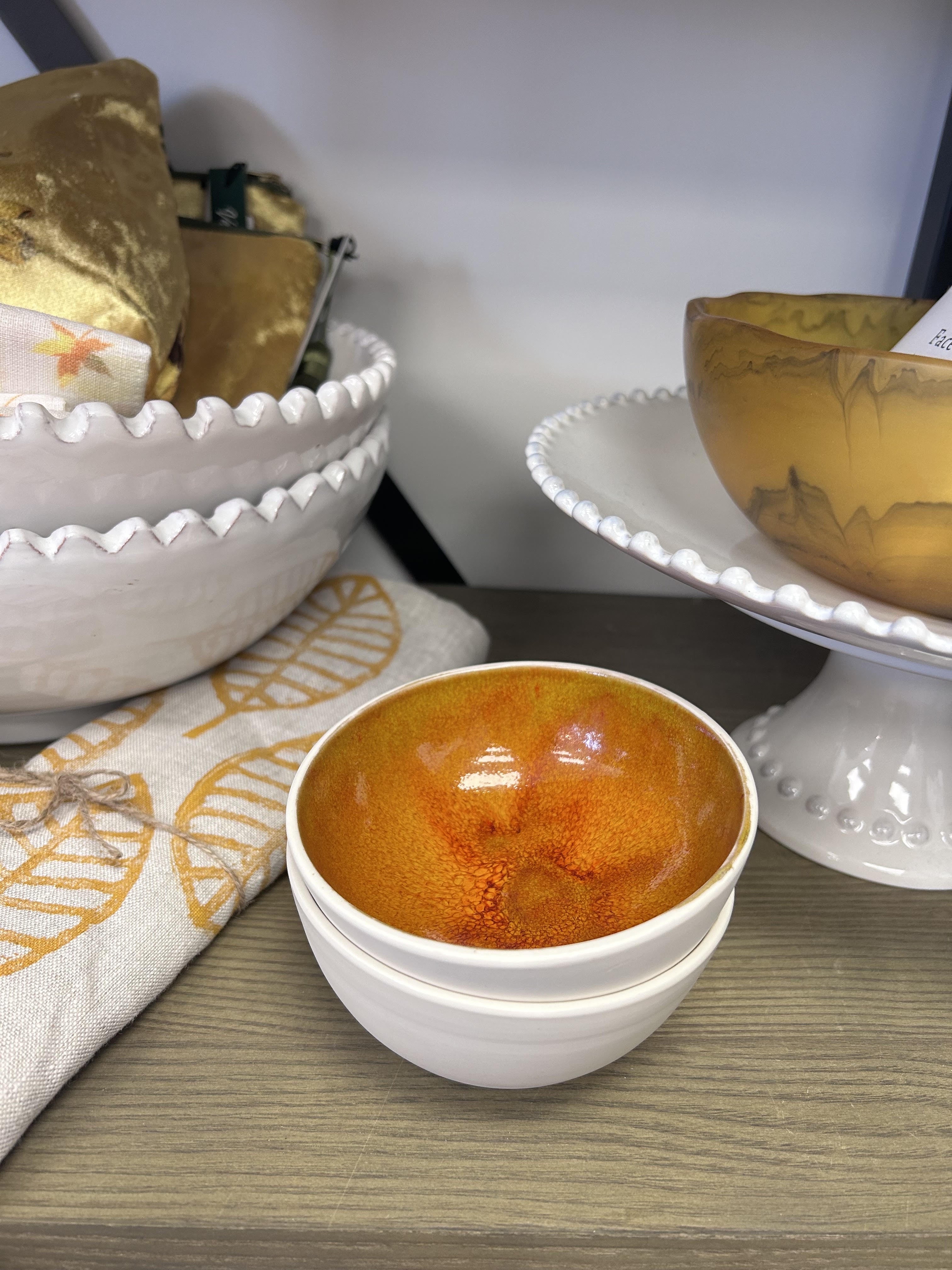A stack of white ceramic bowls with a glossy orange and amber glazed interior, displayed on a wooden surface alongside other decorative bowls and a patterned cloth.