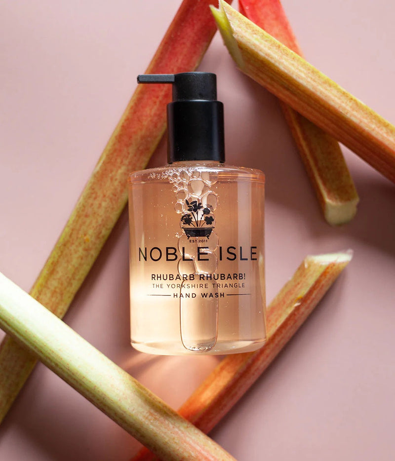 A bottle of Noble Isle Rhubarb & Rhubarbi hand wash with a black pump dispenser, set against a dusty pink background with decorative rhubarb stalks.