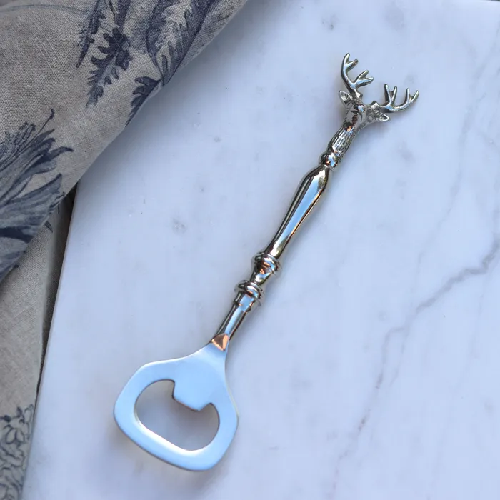 A silver-toned bottle opener shaped like a deer head, placed diagonally on a white marble surface with a patterned fabric in the top-left corner.