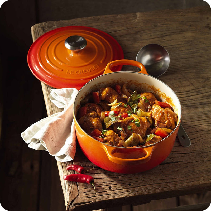 An orange Le Creuset Dutch oven filled with cooked stew rests on a wooden table alongside a ladle, towel, and two red chili peppers—a beautiful scene showcasing the timeless appeal of Le Creuset’s casserole dish.