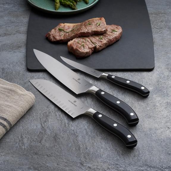 Three Victorinox knives with black handles on a slate board with sliced steak and asparagus