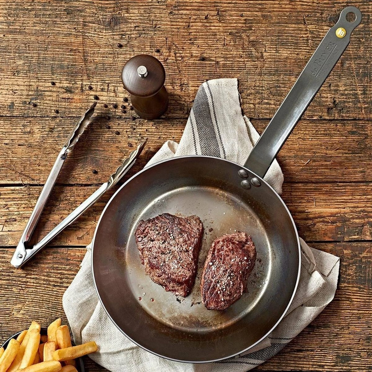 De Buyer carbon steel frying pan searing two steaks, with chips and pepper mill on a wooden table