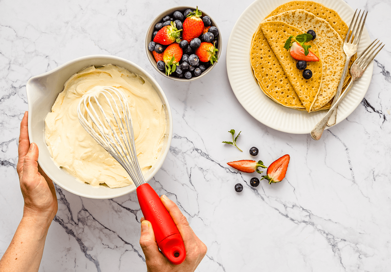 Dreamfarm Flisk red-handled whisk mixing cream in a bowl, with pancakes and berries alongside
