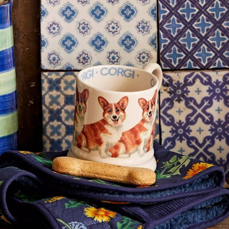A ceramic mug with a hand-painted illustration of Corgi dogs, resting on a navy blue floral pot holder with a biscuit in front, set against a background of patterned blue and white tiles.