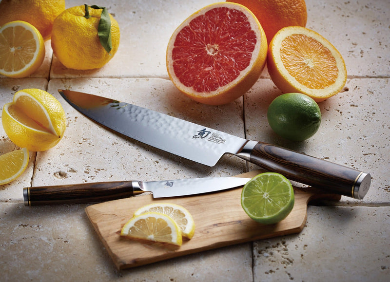 Kai Shun Premier hammered Damascus chef's knife and paring knife with dark wood handles, surrounded by citrus fruit