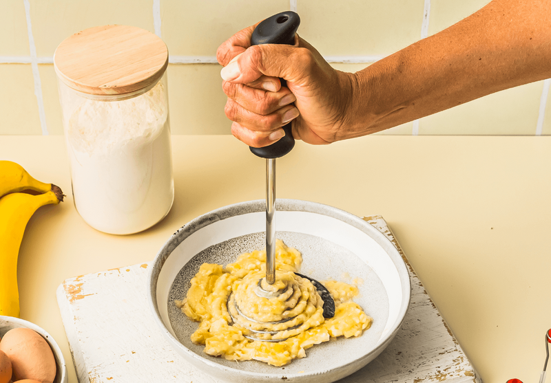 Dreamfarm Smood potato masher mashing ripe bananas in a bowl with flour and eggs nearby