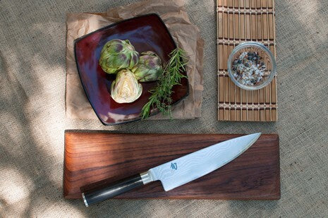 Kai Shun chef's knife with Damascus steel blade resting in a wooden saya, beside a plate of baby artichokes, garlic and herbs
