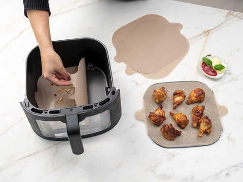 NoStik airfryer liners, a square set of two, shown being placed in an air fryer basket beside cooked chicken