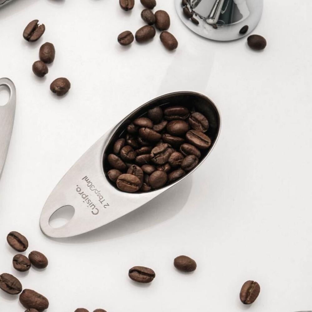 Cuisipro short coffee scoop in stainless steel, filled with coffee beans and surrounded by loose beans