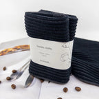 Pack of black barista cloths with a label on a white surface with coffee beans and a spoon.