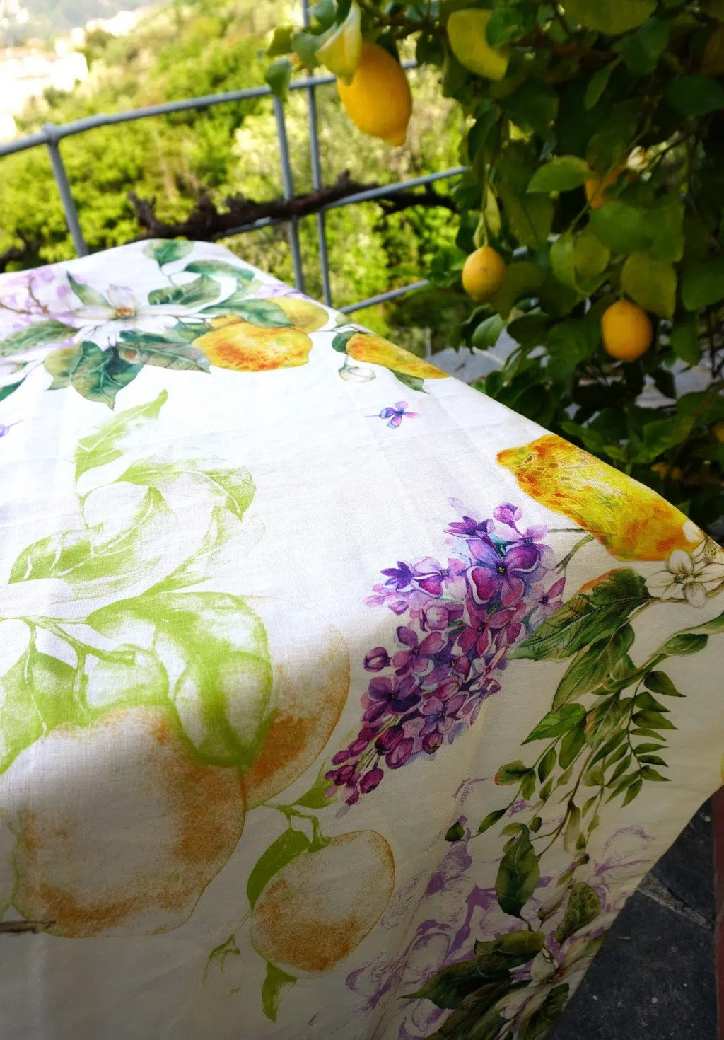 Telene Limoni 100% linen tablecloth with a print of lemons and wisteria, draped over a table under a lemon tree