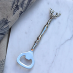 A silver-toned bottle opener shaped like a deer head, placed diagonally on a white marble surface with a patterned fabric in the top-left corner.