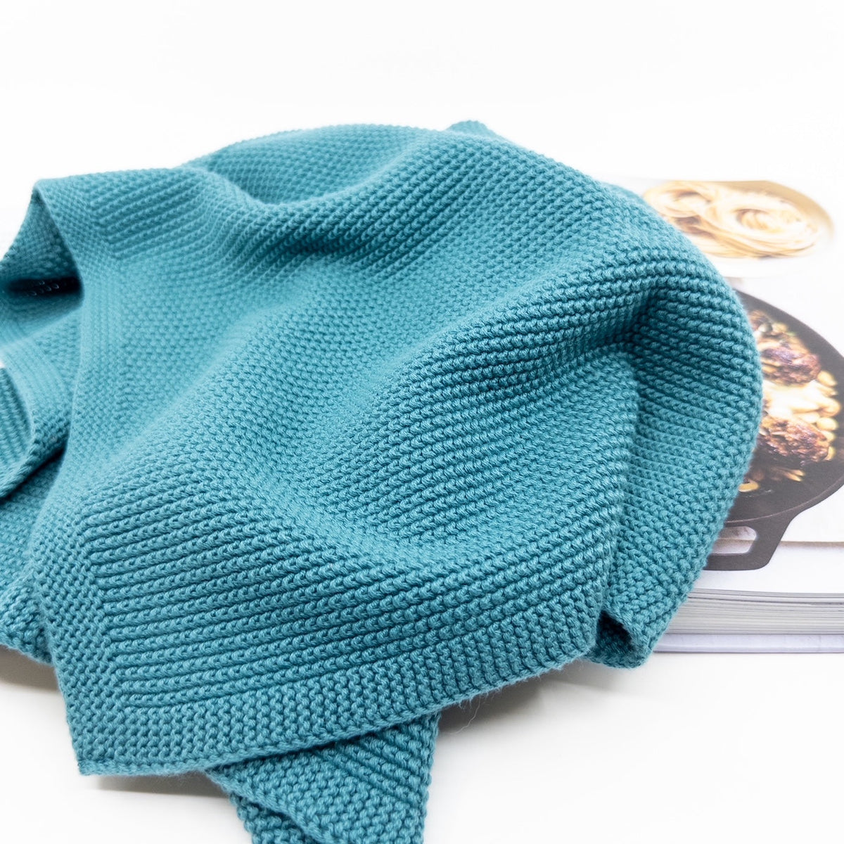 Ecovask hand towel in teal knit, shown folded with a magazine