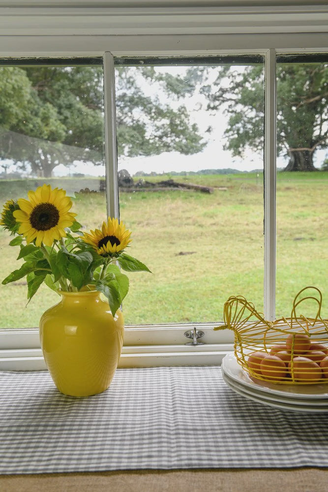 Raine & Humble gingham table runner in ash grey, styled in a window with sunflowers and a wire egg basket