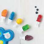 A set of colorful plastic toy popsicle molds and shaped pieces arranged on a light wood surface, including orange, blue, yellow, green, red, and dark purple pieces, with a few blueberries nearby.