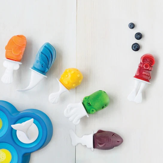 A set of colorful plastic toy popsicle molds and shaped pieces arranged on a light wood surface, including orange, blue, yellow, green, red, and dark purple pieces, with a few blueberries nearby.
