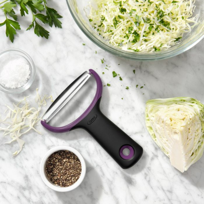 OXO Good Grips large vegetable prep peeler in black with purple trim, shown with shredded cabbage and herbs