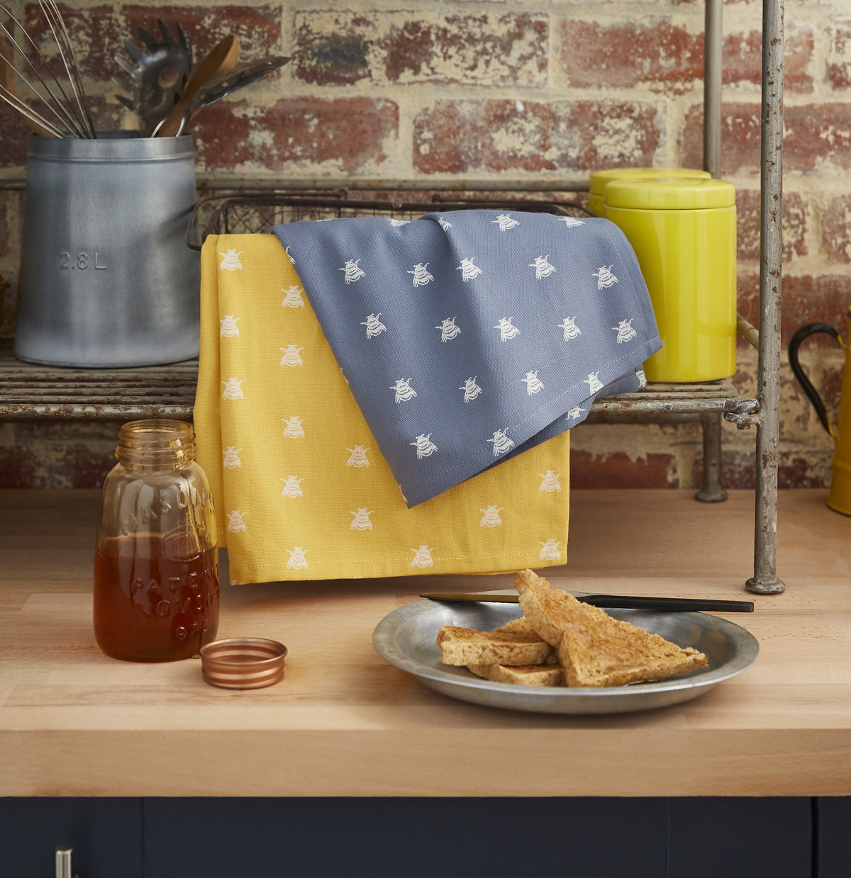 Ulster Weavers cotton tea towel set with bee prints in yellow and blue, styled in a kitchen with toast and honey
