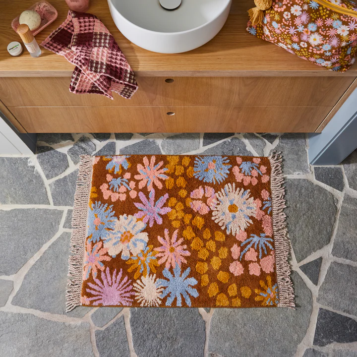Sage x Clare Hayfolk bath mat with a tufted daisy pattern in orange, pink and blue, on a stone bathroom floor
